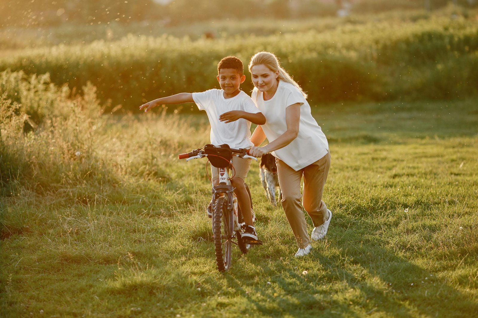 A mother helps her son ride a bicycle on a sunny day in a grassy park.